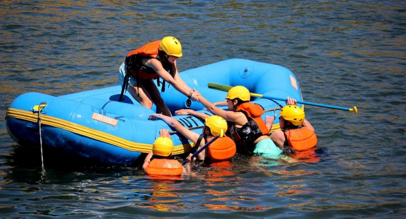 One person in a raft helps others who are in the water into the raft. All are wearing helmets and life jackets.